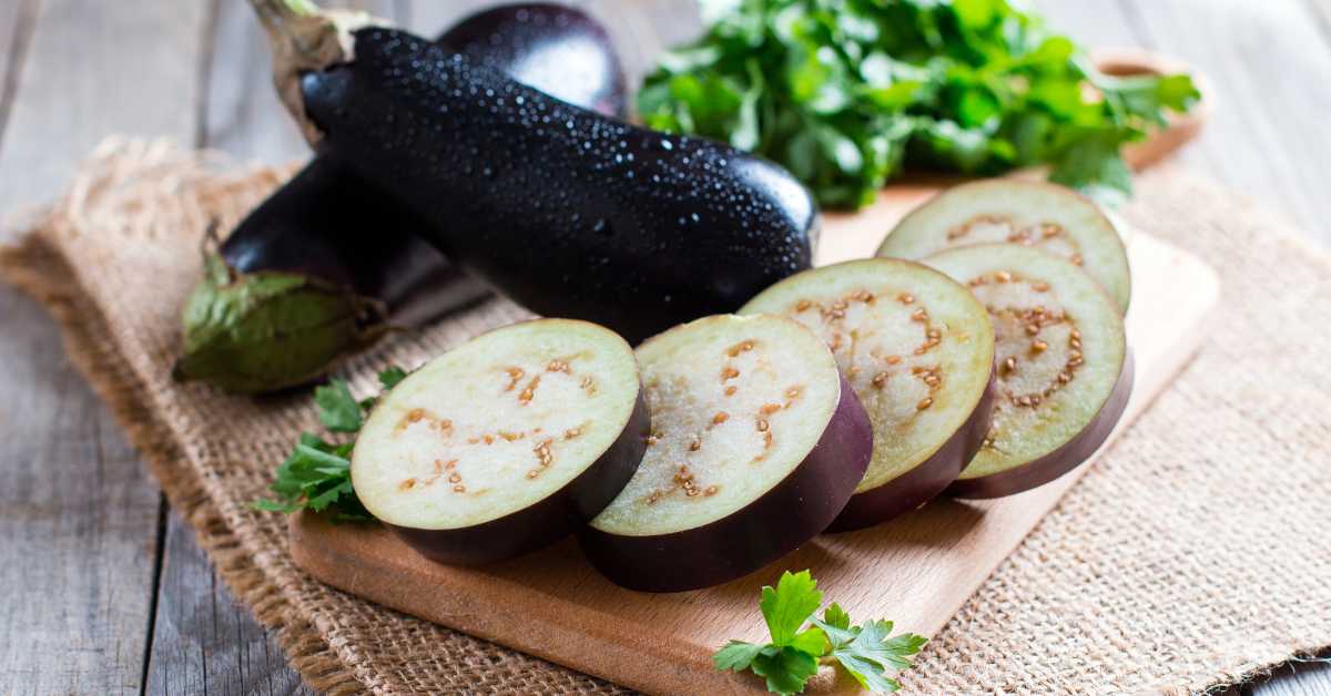 Fresh eggplant sliced on a wooden cutting board with whole eggplants and parsley in the background.